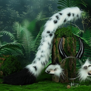 May include: A white and black spotted faux fur tail with a black tip, a black headband with black horns, and a pair of white and black spotted faux fur ears. The items are on a mossy green surface.