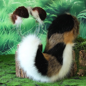 May include: A set of cosplay accessories featuring a fluffy tail and cat ears. The tail is white, black, and brown, and the ears are brown and white. The items are displayed on a mossy log against a green background.