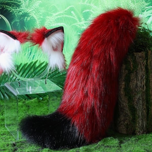 May include: A red and black faux fur tail with a matching headband featuring white tipped ears. The tail is displayed on a clear acrylic stand against a green background.