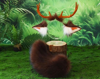 May include: A deer costume set featuring brown antlers, matching ears with white fur, and a fluffy tail. The costume pieces are displayed on a wooden stand against a backdrop of green foliage.