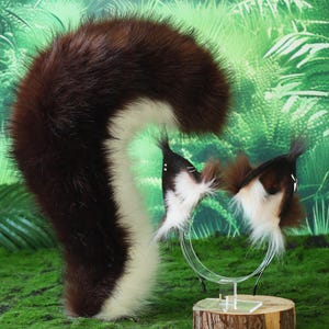 May include: A skunk tail and skunk ear headband set. The tail is brown and white, and the ears are brown, white, and black. The set is displayed on a green mossy surface with a jungle background.