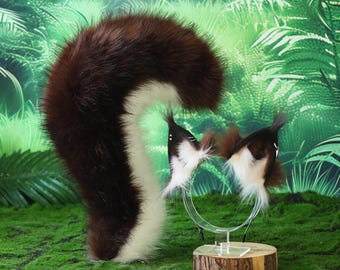 Puede incluir: Un conjunto de cola y diadema de orejas de mofeta. La cola es marrón y blanca, y las orejas son marrones, blancas y negras. El conjunto se exhibe sobre una superficie verde musgosa con un fondo de selva.
