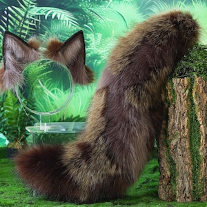 Könnte beinhalten: Ein brauner und schwarzer Kunstfellschwanz und passende Ohren, präsentiert mit einem kleinen Spiegel. Der Schwanz lehnt an einem Baumstumpf mit grünem Moos, vor dem Hintergrund von Farnen und Grün.