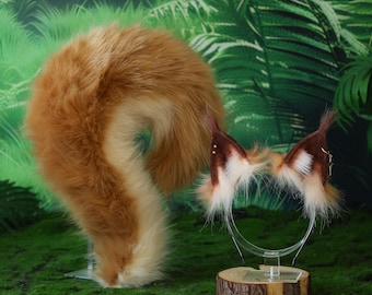 May include: A cosplay set featuring a fluffy, curled fox tail and matching ears on a headband. The tail is a mix of orange and cream-colored faux fur. The ears are brown with white accents and small gold-colored studs. The set is displayed on a green, mossy surface.