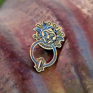 May include: A gold-toned door knocker with a blue and green face and a ring handle. The knocker is on a rusty metal surface.