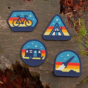 May include: Four embroidered patches with a retro-inspired design. Each patch features a different outdoor activity: a bicycle, a cabin, a camper van, and a rocket. The patches are all navy blue with a sunset background and white stars.