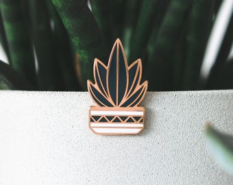 Puede incluir: Un pin de esmalte dorado con una planta en maceta con hojas verde oscuro y una maceta blanca con patrones geométricos negros.