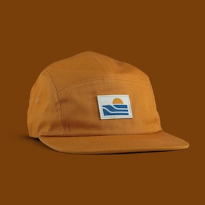 May include: A mustard yellow five-panel cap featuring a rectangular patch on the front. The patch displays a graphic of a yellow sun rising over blue wavy lines against a white background. The cap has a flat brim and a structured crown.