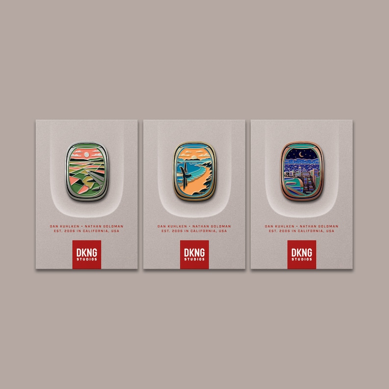 May include: Three enamel pins with different landscape designs. Each pin features a stylized image of a sunset, a beach with a palm tree, and a cityscape with a crescent moon. The pins are all rectangular and have a white background with black text that reads "DKNG Studios".