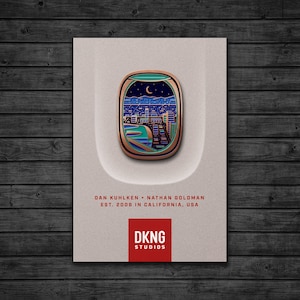 May include: A gold and blue enamel pin featuring a cityscape seen through an airplane window. The pin has a green background with a crescent moon and stars. The text "DKNG STUDIOS" is printed in red below the pin.