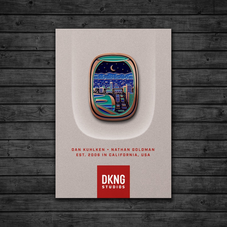 May include: A gold and blue enamel pin featuring a cityscape seen through an airplane window. The pin has a green background with a crescent moon and stars. The text "DKNG STUDIOS" is printed in red below the pin.