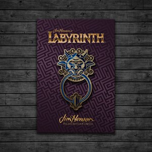 May include: A gold and blue enamel pin featuring a goblin-like face with a large ring, inspired by the movie Labyrinth. The pin is on a purple background with a maze pattern and the text "Jim Henson's Labyrinth" and "Jim Henson The Jim Henson Company".