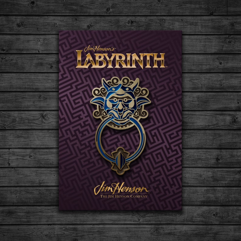 May include: A gold and blue enamel pin featuring a goblin-like face with a large ring, inspired by the movie Labyrinth. The pin is on a purple background with a maze pattern and the text "Jim Henson's Labyrinth" and "Jim Henson The Jim Henson Company".