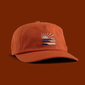 May include: An orange baseball cap with a low profile. The cap features an embroidered design of a sun over blue and white horizontal lines. The hat is made of a soft, durable fabric and has a curved peak.