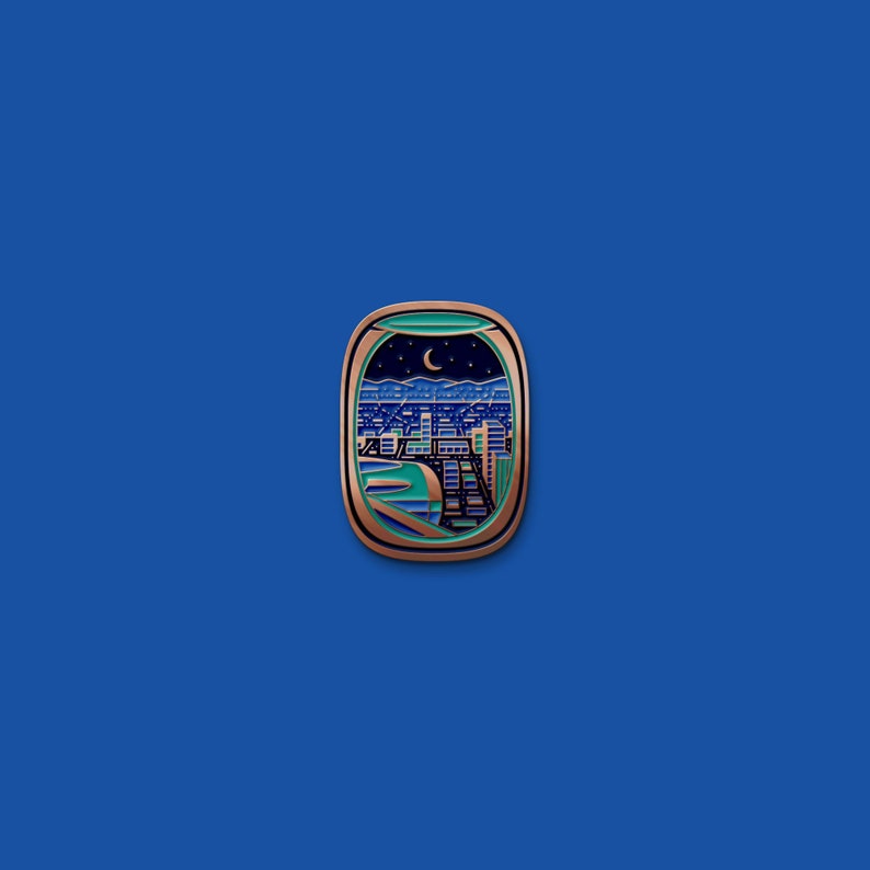 May include: A copper-colored airplane window pin with a blue and green enamel cityscape view. The cityscape is lit up at night with a crescent moon in the sky.