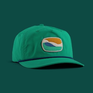 Puede incluir: Gorra de béisbol verde con visera curva y detalle de cuerda azul. La gorra presenta un parche rectangular con un diseño de paisaje bordado en naranja, azul y verde. La gorra es de color verde liso.