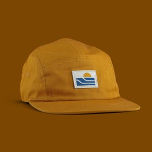 May include: Mustard-colored five-panel cap with a flat brim. A square patch on the front features a blue and white ocean scene with a yellow sun. The cap is made of a durable fabric and has a classic, casual style.