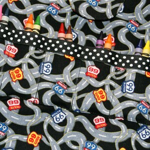 May include: A black fabric apron with a white and black polka dot ribbon. The apron has multiple pockets with the words "Route 66" and "Route 99" printed on them. The pockets are filled with crayons.