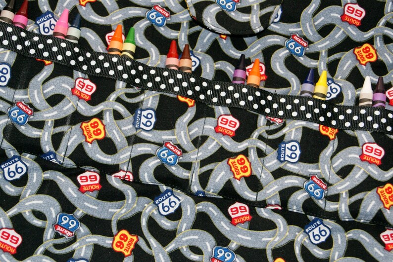 May include: A black fabric apron with a white and black polka dot ribbon. The apron has multiple pockets with the words "Route 66" and "Route 99" printed on them. The pockets are filled with crayons.