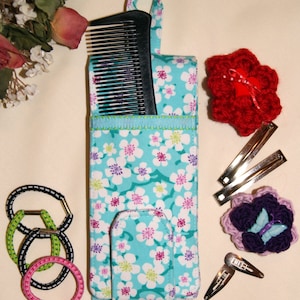 May include: A teal floral print hair accessory organizer with a black comb, hair clips, and hair ties. The organizer has a handle and a flap. Crochet flower hair accessories in red and purple are also visible.