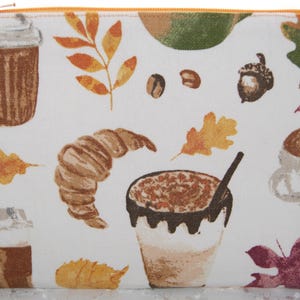 May include: A white zippered pouch featuring a fall-themed pattern. The design includes illustrations of coffee drinks, croissants, acorns, coffee beans, and autumn leaves in shades of brown, orange, and green.