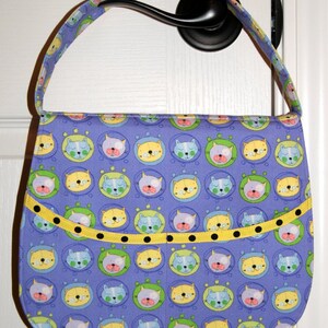 May include: A purple fabric tote bag with a yellow and black trim. The bag is decorated with a repeating pattern of cartoon cats in bubbles.