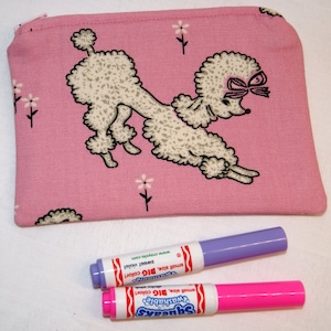 May include: Pink fabric zippered pouch with a white poodle design. The pouch is decorated with small white flowers. The pouch is 7 inches wide and 4 inches tall.