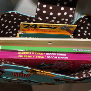 May include: A brown and white polka dot tote bag with a blue and white lining. The bag is filled with three books, including a green book titled "Super WilY! Jumbo Activity Book" and two pink books titled "Minnie Mouse - What a SweetIe!"  The bag also contains a yellow box of 24 crayons.