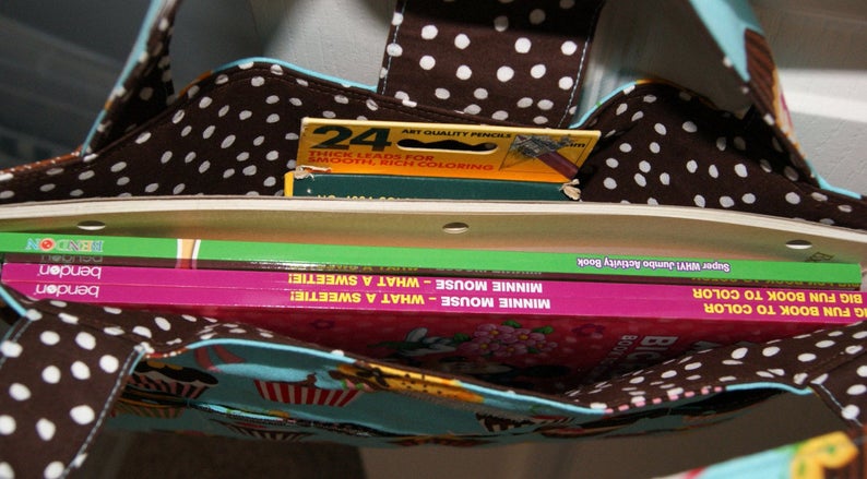 May include: A brown and white polka dot tote bag with a blue and white lining. The bag is filled with three books, including a green book titled "Super WilY! Jumbo Activity Book" and two pink books titled "Minnie Mouse - What a SweetIe!"  The bag also contains a yellow box of 24 crayons.