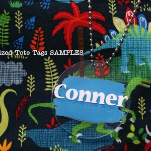 May include: A round, clear plastic tote tag with a blue background and white lettering that reads "Conner". The tag is attached to a silver chain and is hanging from a black fabric tote bag with a dinosaur print.
