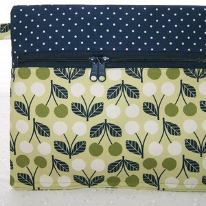 Può includere: Una pochette in tessuto verde e bianco con chiusura a zip. La pochette presenta un motivo ripetitivo di ciliegie e foglie. La parte superiore della pochette è blu navy con pois bianchi.