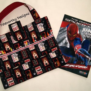 May include: A black and red tote bag with a dog-themed print and a pocket for crayons. The bag has a red and white striped handle. The bag is next to a coloring book featuring The Amazing Spider-Man. The coloring book has a red, blue, and black cover with the title "The Amazing Spider-Man Jumbo Coloring and Activity Book".