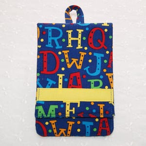 May include: A blue fabric notepad cover with a colourful alphabet pattern. The cover has a yellow ribbon accent and a loop at the top. The letters are in various colours, including red, green, and blue.