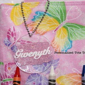 May include: A pink tote bag with a butterfly pattern and a clear plastic tag with the name "Gwyneth" written in white letters. The tag is attached to the bag with a silver chain. The text "Personalized Tote Tags SAMPLES" is printed on the bag.