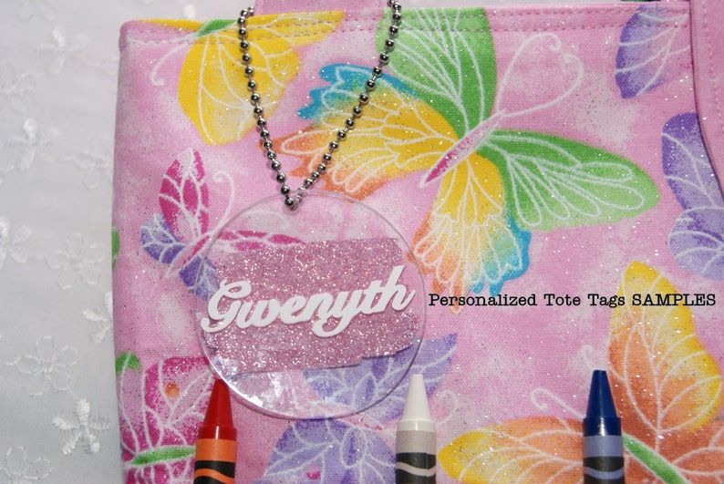 May include: A pink tote bag with a butterfly pattern and a clear plastic tag with the name "Gwyneth" written in white letters. The tag is attached to the bag with a silver chain. The text "Personalized Tote Tags SAMPLES" is printed on the bag.