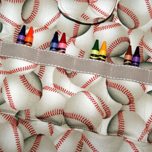 May include: A gray fabric pouch with a white and red baseball pattern. The pouch has a gray ribbon across the top with a row of colored crayons inserted into the ribbon.