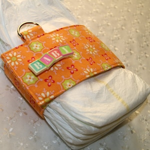 May include: Orange and green fabric diaper holder with a white plastic "BABY" sign. The holder is attached to a stack of white diapers.