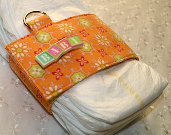 Puede incluir: Portapañales de tela naranja y verde con un letrero de plástico blanco que dice "BABY". El portapañales está sujeto a una pila de pañales blancos.