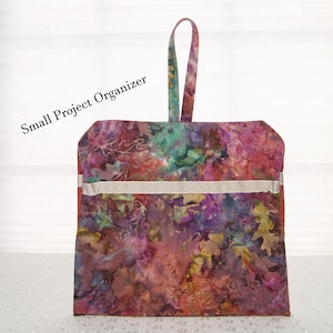 May include: A small project organizer bag with a handle. The bag is made of a colorful fabric with a floral pattern in shades of pink, purple, green, and brown. The bag has a white ribbon trim.