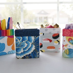 May include: Four fabric storage containers with different patterns. The containers are filled with various items, including pens, pencils, candy, and toiletries. The containers are all square and have a white background with colorful patterns.