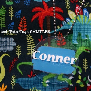 May include: A round, clear plastic tote tag with a blue background and white lettering that reads "Conner". The tag is attached to a silver chain and is hanging from a black fabric tote bag with a dinosaur print.