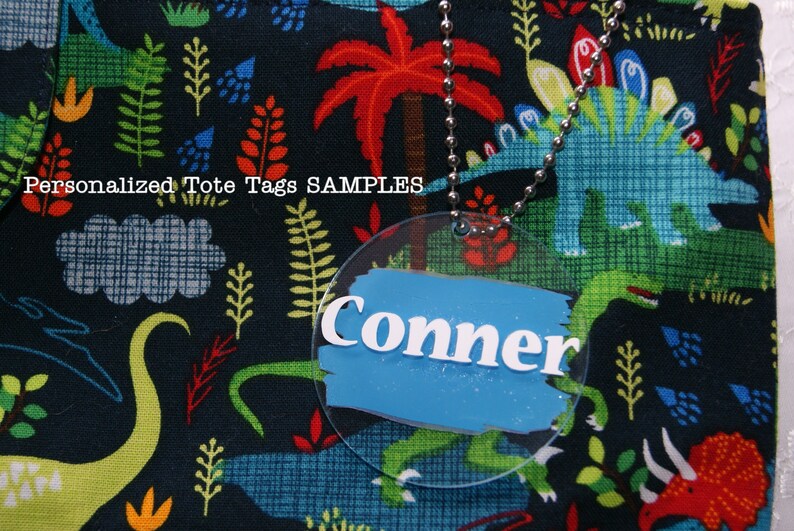 May include: A round, clear plastic tote tag with a blue background and white lettering that reads "Conner". The tag is attached to a silver chain and is hanging from a black fabric tote bag with a dinosaur print.