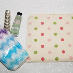 May include: A white polka dot makeup bag with pink and green polka dots. The bag is open and contains a black eyeglass case, a small tube of hand cream, a package of tissues, and a silver hair roller.