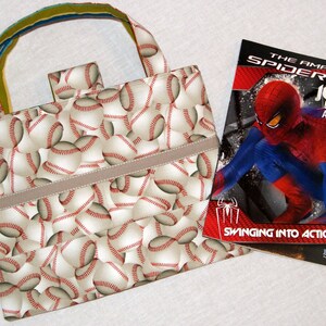 May include: A beige tote bag with a baseball pattern and a brown strap. The bag is next to a coloring book featuring Spiderman swinging into action. The book cover is red, blue, and black with the title "The Amazing Spider-Man Jumbo Coloring and Activity Book".