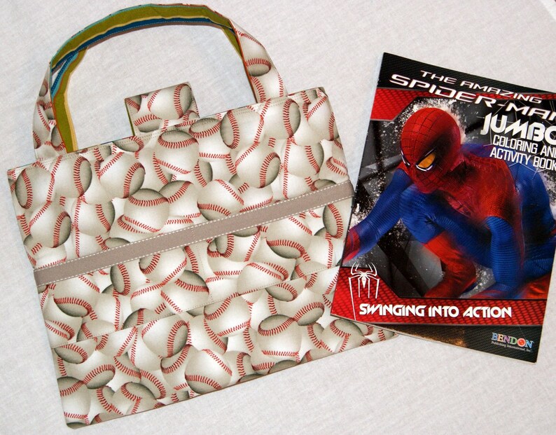 May include: A beige tote bag with a baseball pattern and a brown strap. The bag is next to a coloring book featuring Spiderman swinging into action. The book cover is red, blue, and black with the title "The Amazing Spider-Man Jumbo Coloring and Activity Book".