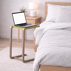 Może przedstawiać: Laptop z pustym ekranem stoi na małym stoliku. Stół ma jasnozielony blat i zakrzywioną, neutralną ramę. Scena rozgrywa się w sypialni z łóżkiem pokrytym białą pościelą.