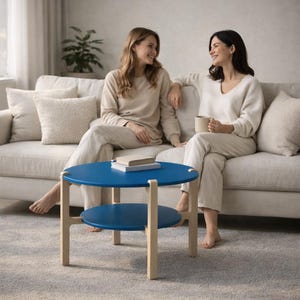 Könnte beinhalten: Ein zweistufiger Couchtisch mit blauer Tischplatte und hellem Holzrahmen. Der Tisch hat eine runde Oberseite und ein unteres Regal. Der Tisch steht auf einem grauen Teppich. Im Hintergrund sitzen zwei Personen auf einem beigen Sofa.