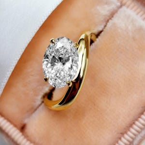 May include: A gold engagement ring featuring a large, oval-cut diamond. The ring is set in a classic four-prong setting and is presented in a pink velvet ring box. The band has a smooth, polished finish.