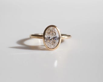 May include: A gold ring with an oval-shaped diamond. The diamond is set in a bezel setting, and the band is thin and delicate. The ring is on a white surface, and the lighting creates a shadow.
