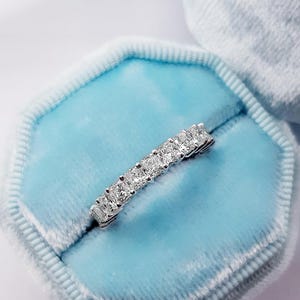 May include: A silver ring with a row of square-cut diamonds, set in a band. The ring is displayed in a light blue velvet ring box. The diamonds sparkle, reflecting light. The ring is a piece of jewelry.